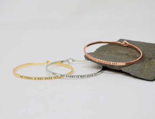 Inspirational "My Story is Not Over Yet" stackable bangle bracelets in gold, silver, and rose gold on a stone surface