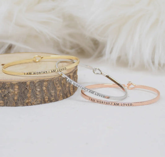 Inspirational "I am Worthy, I am Loved" stackable bangle bracelets in gold, silver, and rose gold on a wooden surface.